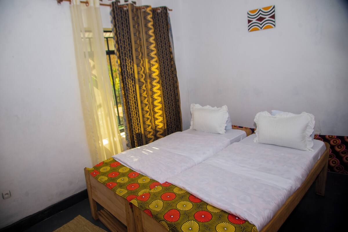 A double bed with a patterned bedspread, two pillows, a wooden headboard, and a curtain with a zigzag pattern.