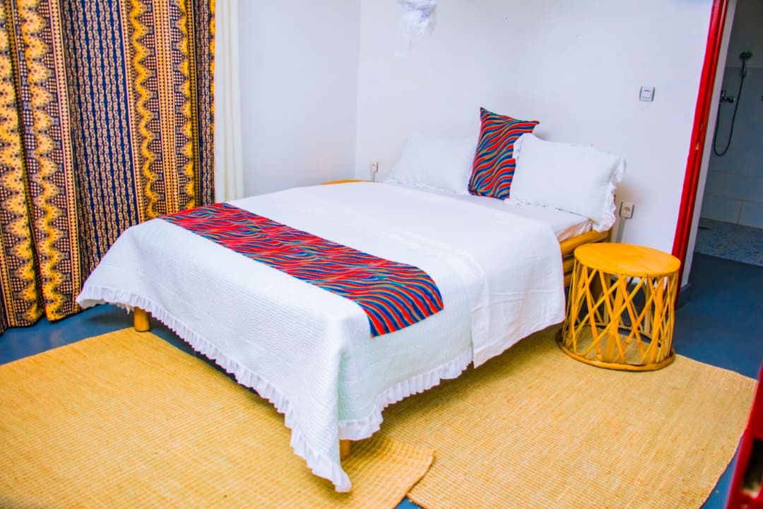 A single bed with a white bedspread and a colorful striped blanket, a wooden stool, and a patterned curtain.