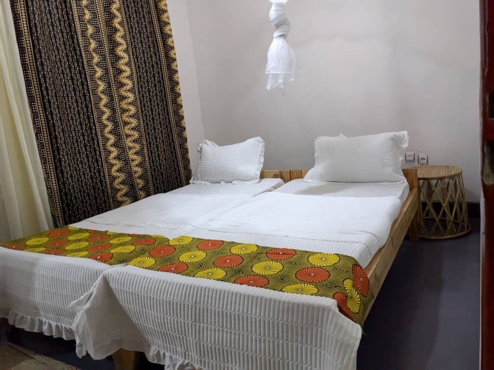 Una cama doble con ropa blanca de cama y una colcha floral colorida. Hay dos almohadas blancas y una pequeña mesa redonda con base trenzada.