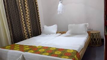 Una cama doble con ropa blanca de cama y una colcha floral colorida. Hay dos almohadas blancas y una pequeña mesa redonda con base trenzada.
