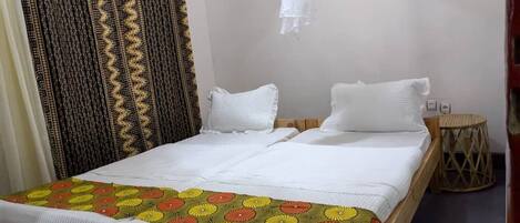 A double bed with white bedding and a colorful floral bedspread. There are two white pillows and a small round table with a woven base.