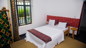 A neatly made bed with a patterned bedspread, a bedside table with a lamp, and a window with a view of greenery. - Ineza Guest House (Kinoni)