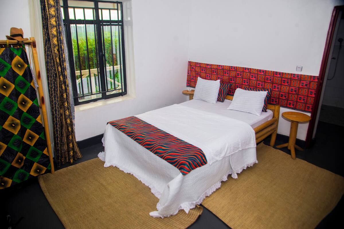 A neatly made bed with a patterned bedspread, a bedside table with a lamp, and a window with a view of greenery.