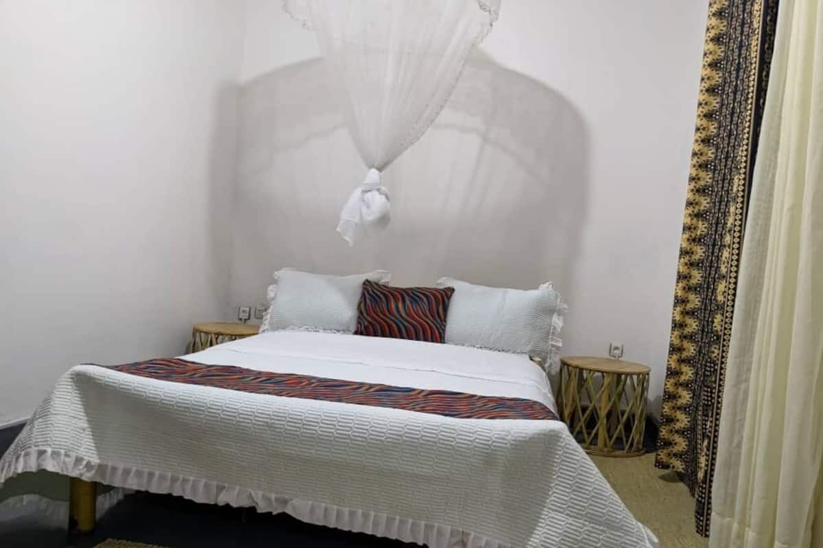 A neatly made bed with a white bedspread and a colorful pillow. There is a small round table beside the bed and a curtain with a patterned design.