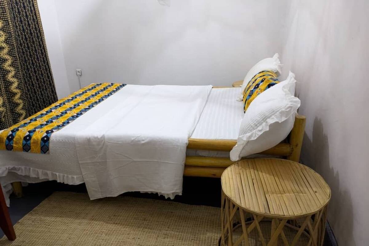 A single bed with a white bedspread and a patterned blanket, a wooden stool, and a woven mat on the floor.