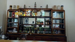A bar area with shelves of bottles and glasses, a wooden cabinet with decorative items, and a person seated at a table. - Ineza Guest House (Kinoni)