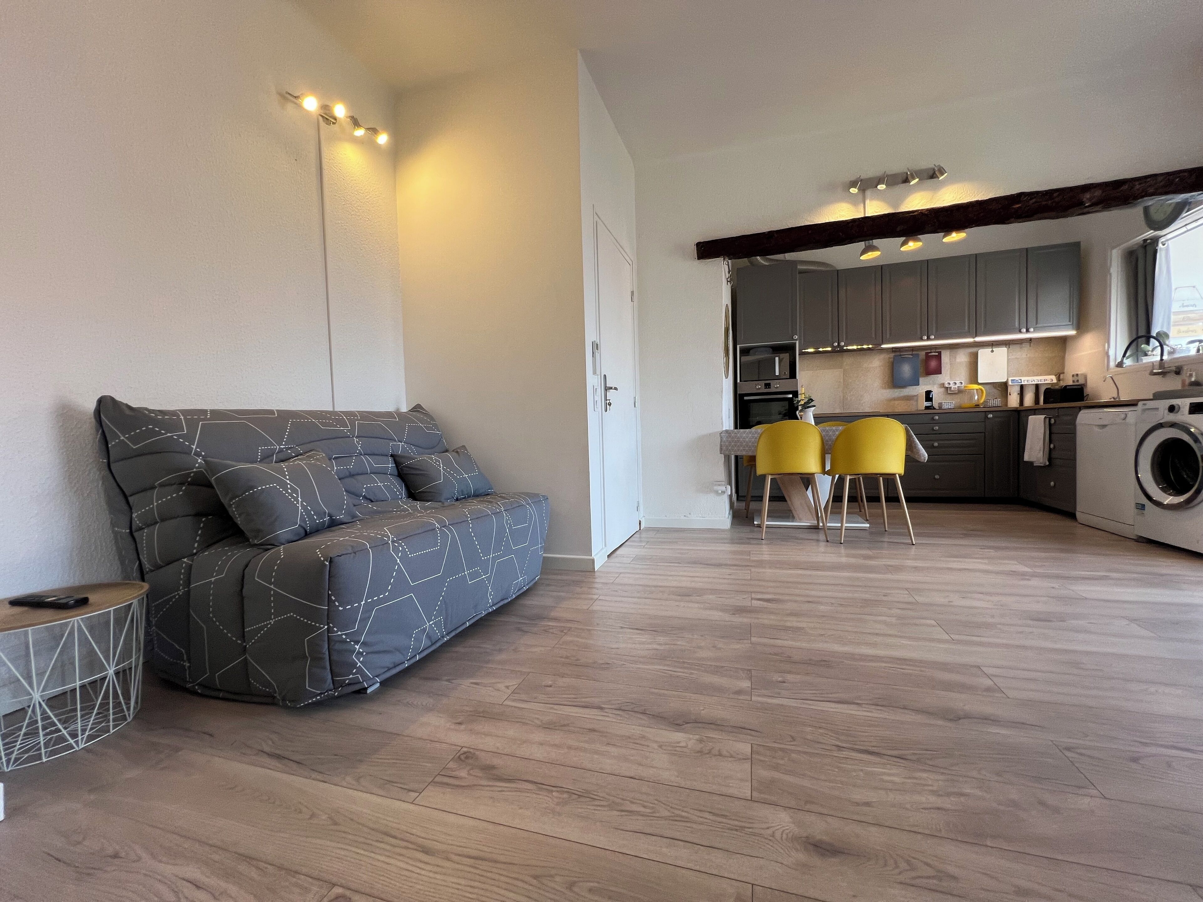 Una sala moderna con un sofá gris, suelo de madera y una zona de cocina con lavadora.