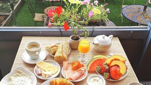 Un balcón con un desayuno que incluye huevos, tostadas, bollería y fruta, con vistas a un jardín con mesas y sillas.