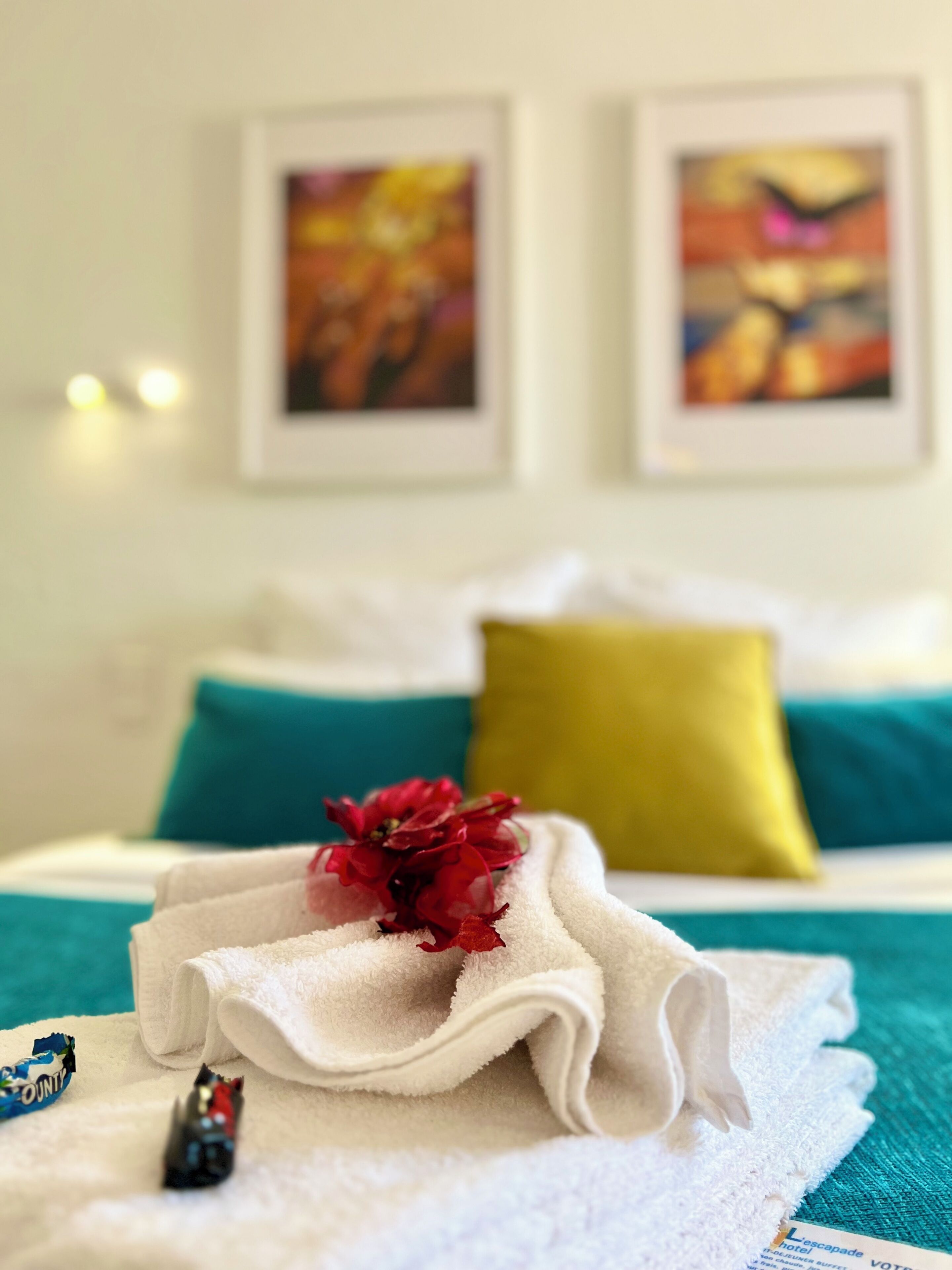 Un lit bien fait, avec une serviette blanche, une fleur rouge et une petite figurine. Il y a deux oreillers, un vert et un jaune, et deux œuvres d’art encadrées accrochées au mur.