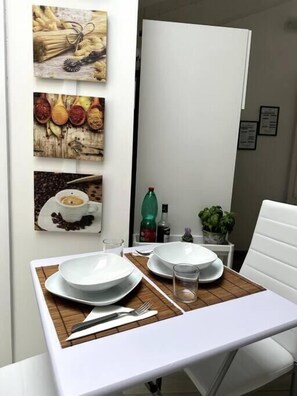 En matplats med ett vitt bord dukat för två personer, en grön flaska och en krukväxt på ett litet bord.