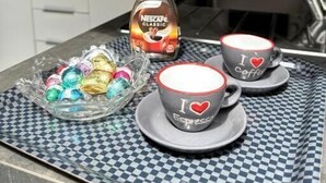 Una encimera de cocina con una bandeja de cuadros que contiene macarons de colores, una botella de Nescafé Classic y dos tazas de espresso con la inscripción 'I love coffee'.
