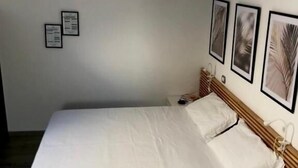 Una cama individual con una colcha y almohadas blancas, una mesita de noche con una taza y un marco de fotos, y tres cuadros con motivos de hojas en la pared.
