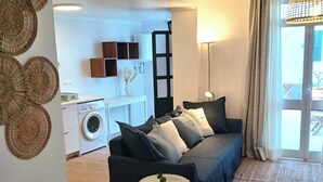 A modern living room with a sofa, a small side table, and a woven rug. There is a washing machine and a kitchen area in the background. - SandyBeachApartment3 (Porto Santo)
