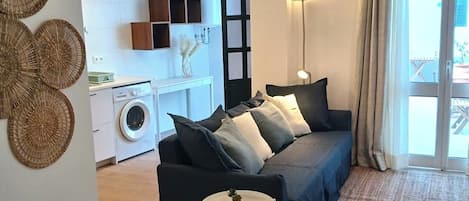 A modern living room with a sofa, a small side table, and a woven rug. There is a washing machine and a kitchen area in the background.