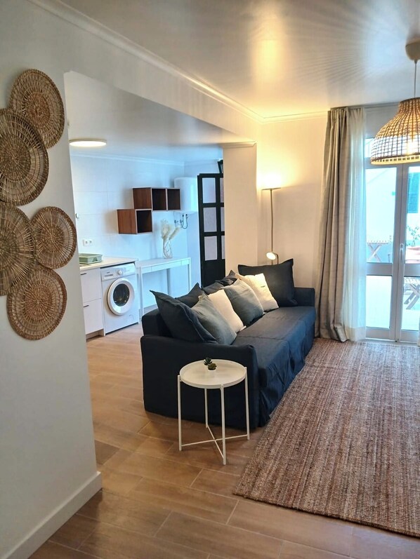 Un salon moderne avec un canapé, une petite table d’appoint et un tapis tissé. On aperçoit une machine à laver et un coin cuisine au fond de la pièce.