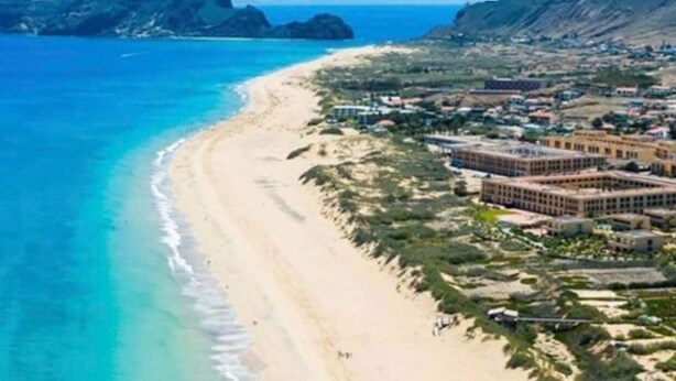An aerial view of a coastal area with a sandy beach, a clear blue sea, and a cluster of buildings with a flat roof design. - SandyBeachApartment3 (Porto Santo)