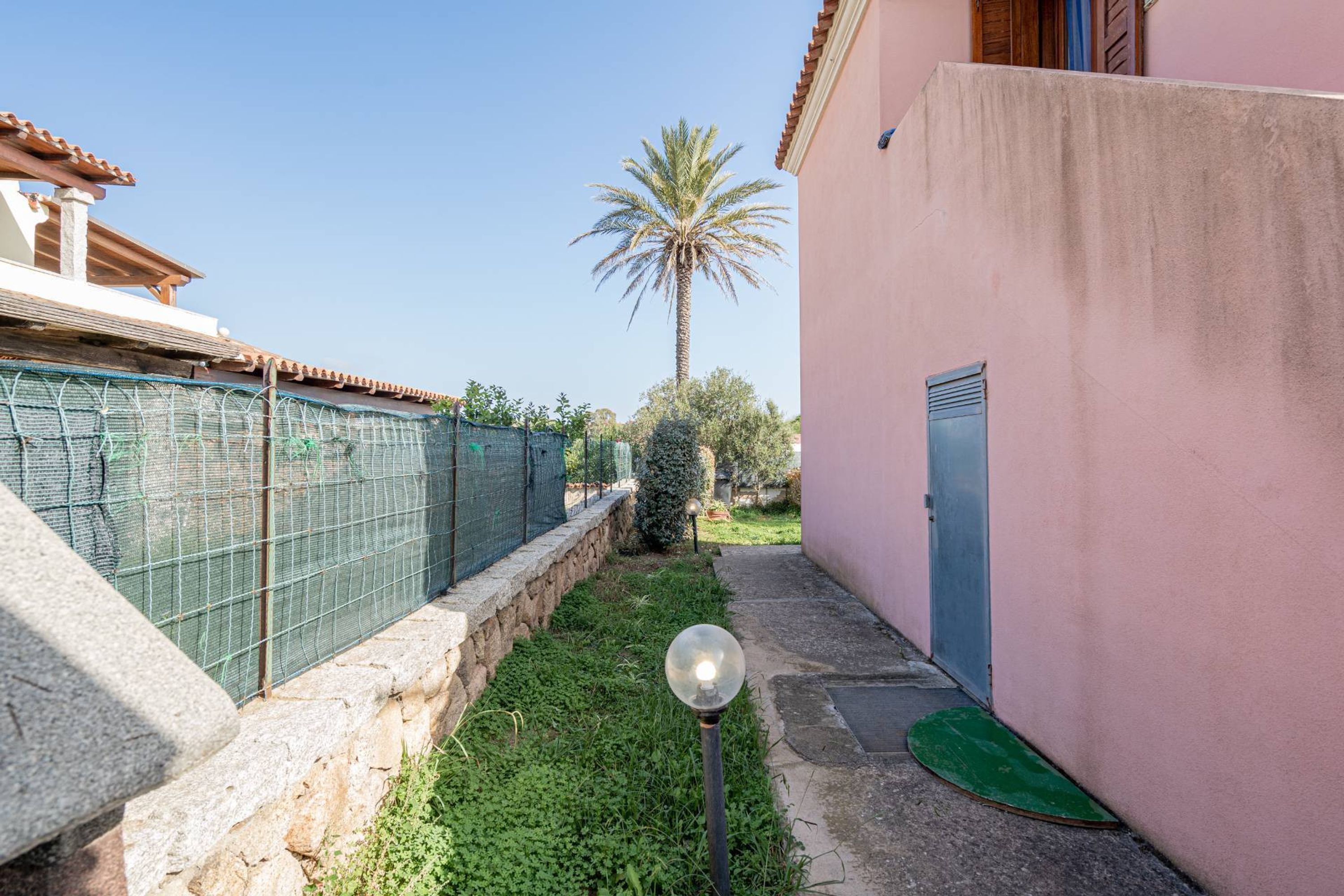 A narrow alley with a stone wall, a green fence, a palm tree, and a building with a pink wall and a blue door.