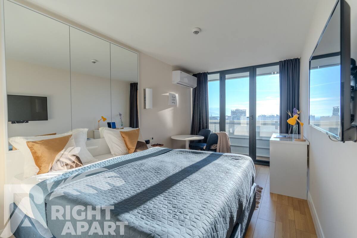 A modern hotel room with a large bed, a small desk, a chair, a TV, and a city view through the windows.