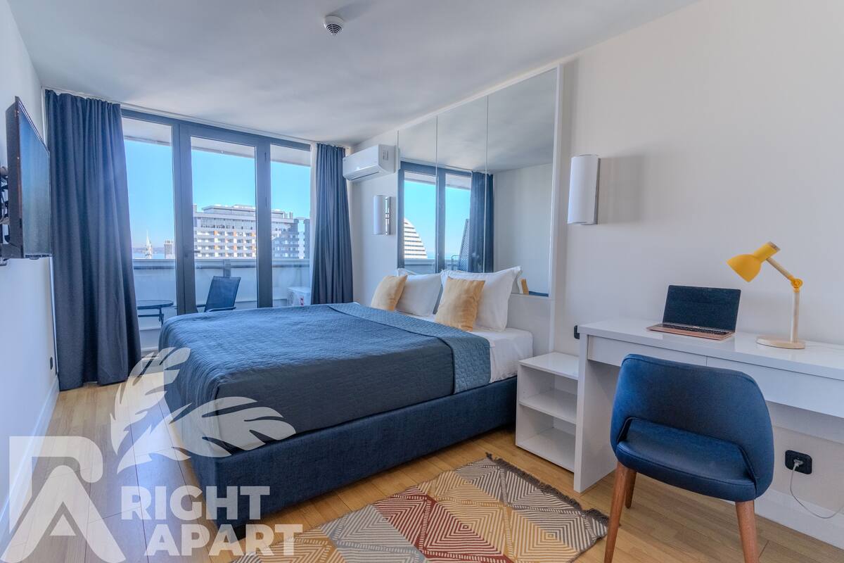 A modern bedroom with a bed, a desk, and a chair. There is a window with curtains, a laptop on the desk, and a view of buildings outside.