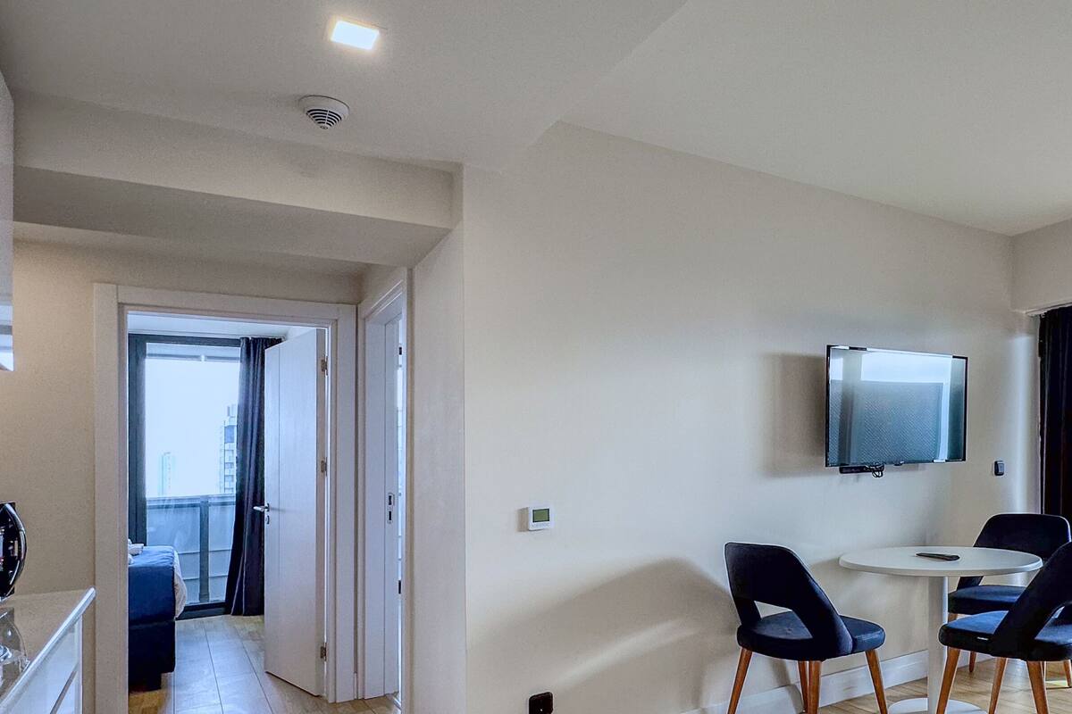 A modern apartment with a balcony, a dining area with two chairs, a TV mounted on the wall, and a kitchen area.