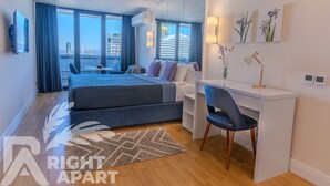 A modern hotel room with a bed, desk, chair, and large windows offering a city view. - ORBI CITY RightApart Hotel (Batumi)