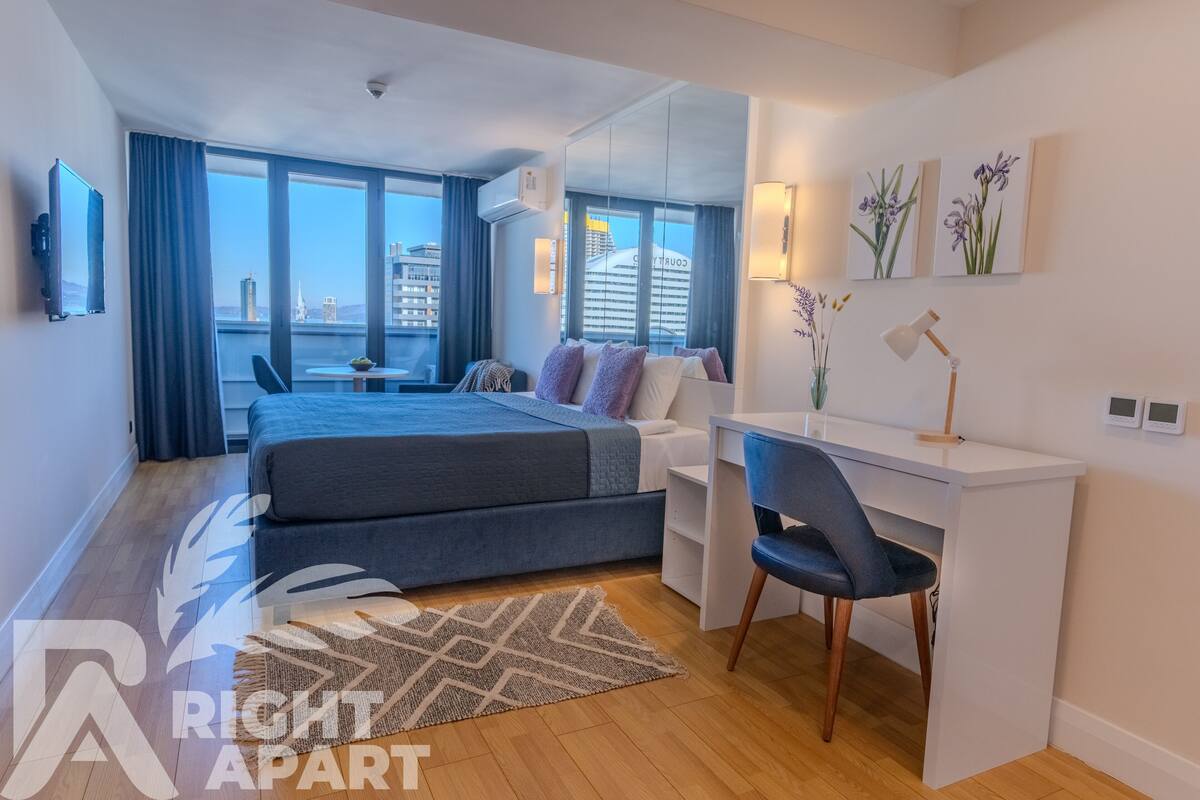 A modern hotel room with a bed, desk, chair, and large windows offering a city view.