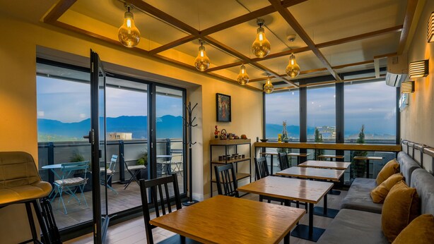 A restaurant with wooden tables and chairs, a sofa, and a view of mountains through large windows. - ORBI CITY RightApart Hotel (Batumi)