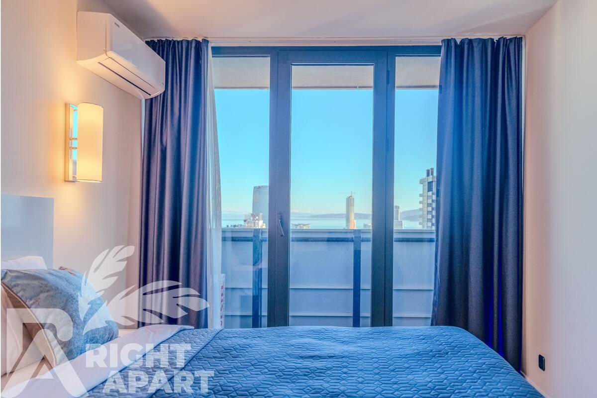 A hotel room with a bed, an air conditioning unit, a wall-mounted light, and a view of the city through large windows.