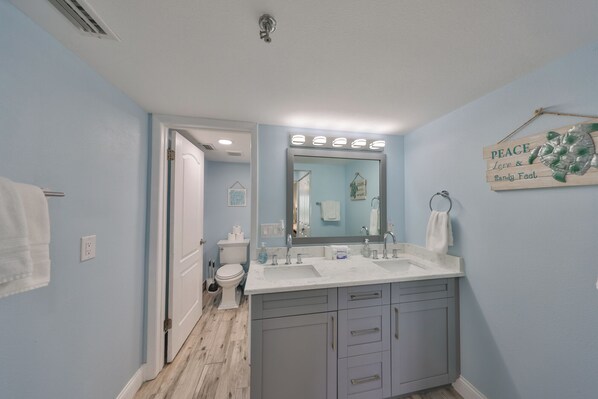 A bathroom with a large mirror, two sinks, and a toilet. There is a towel rack and a sign on the wall.