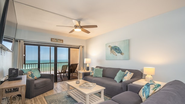 A living room with a sofa, coffee table, and a TV. There is a balcony with a view of the ocean.