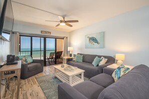 A living room with a sofa, coffee table, and a TV. There is a balcony with a view of the ocean.