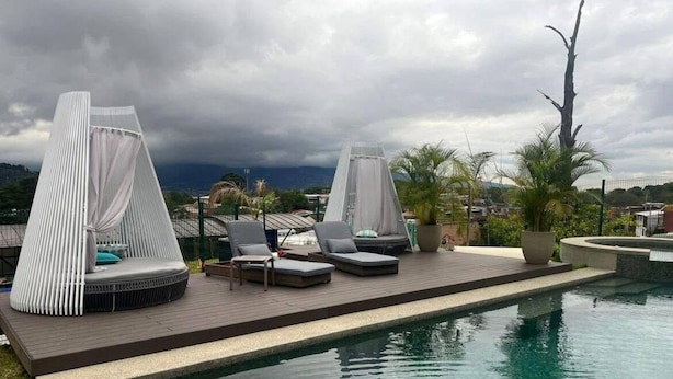 Zona de piscina con sillones de descanso y un área de estar cubierta, rodeada por una piscina y vistas a la ciudad.