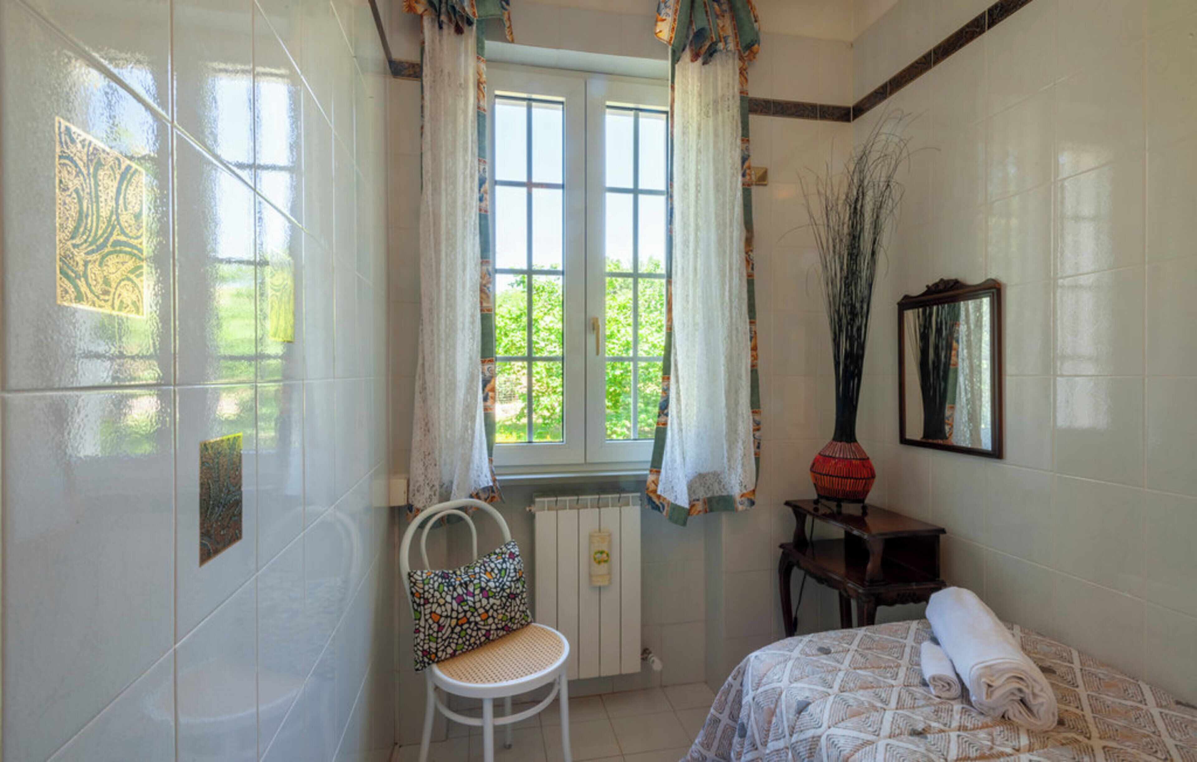 Un baño con ventana, un radiador, una silla, una cama con una toalla enrollada y un florero sobre una mesa.