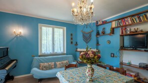 A living room with a piano, a blue sofa, a dining table with a floral tablecloth, and a chandelier. - VILLA WITH POOL FOR 14 PEOPLE COMPLETELY FENCED IDEAL FOR VISITING (Marche)