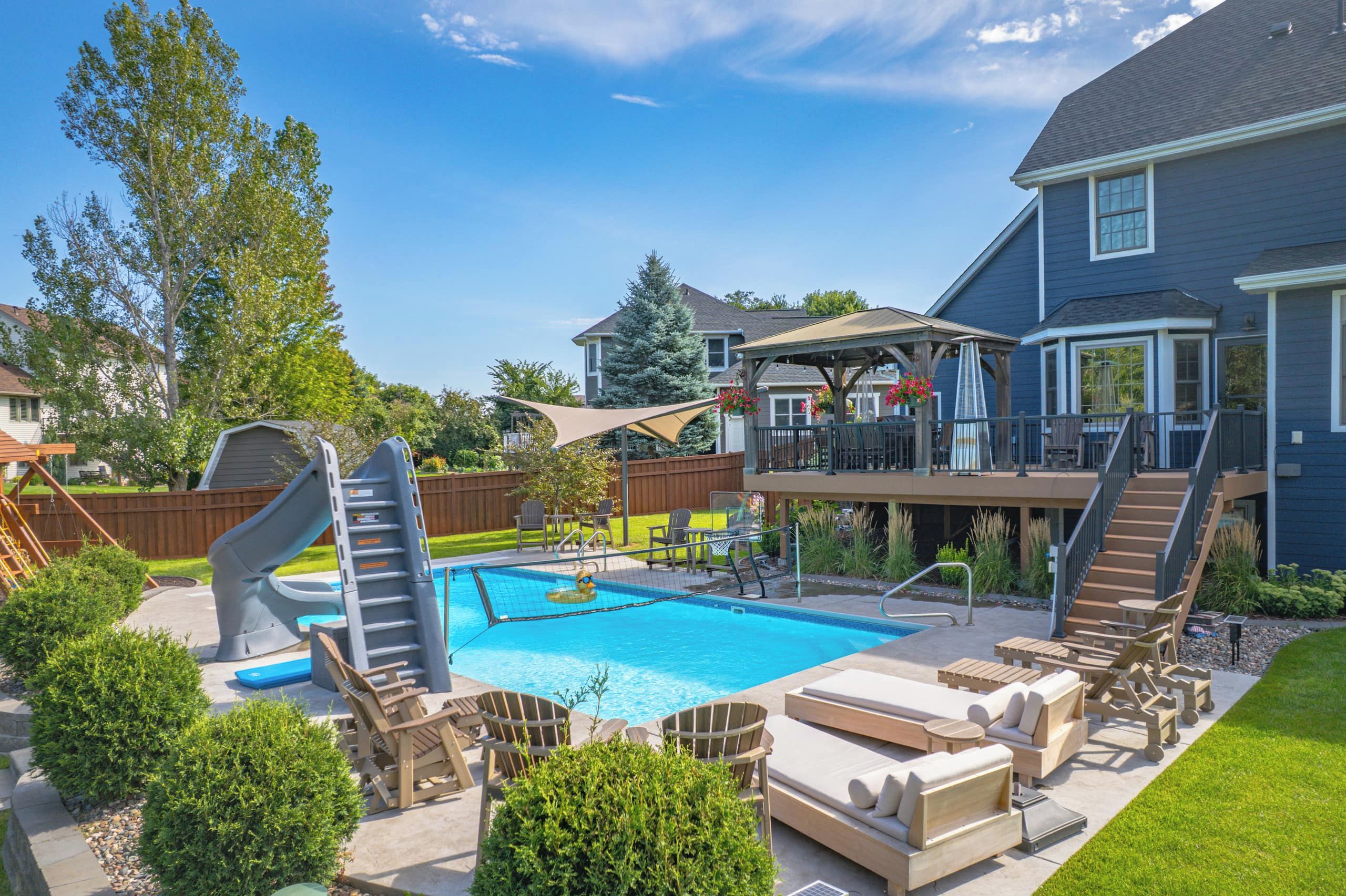 A backyard with a swimming pool, a slide, lounge chairs, and a deck with outdoor furniture.