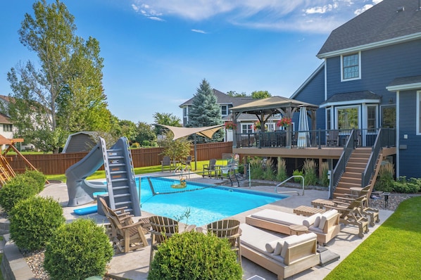 A backyard with a swimming pool, a slide, lounge chairs, and a deck with outdoor furniture.