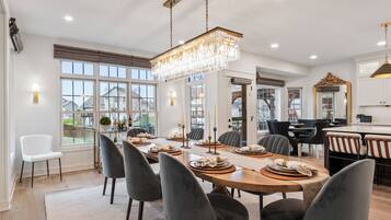 Une salle à manger moderne, avec une grande table en bois dressée pour six personnes, entourée de chaises aux couleurs sombres. Un lustre est suspendu au plafond, et la pièce est dotée de grandes fenêtres qui offrent une vue sur l’extérieur.
