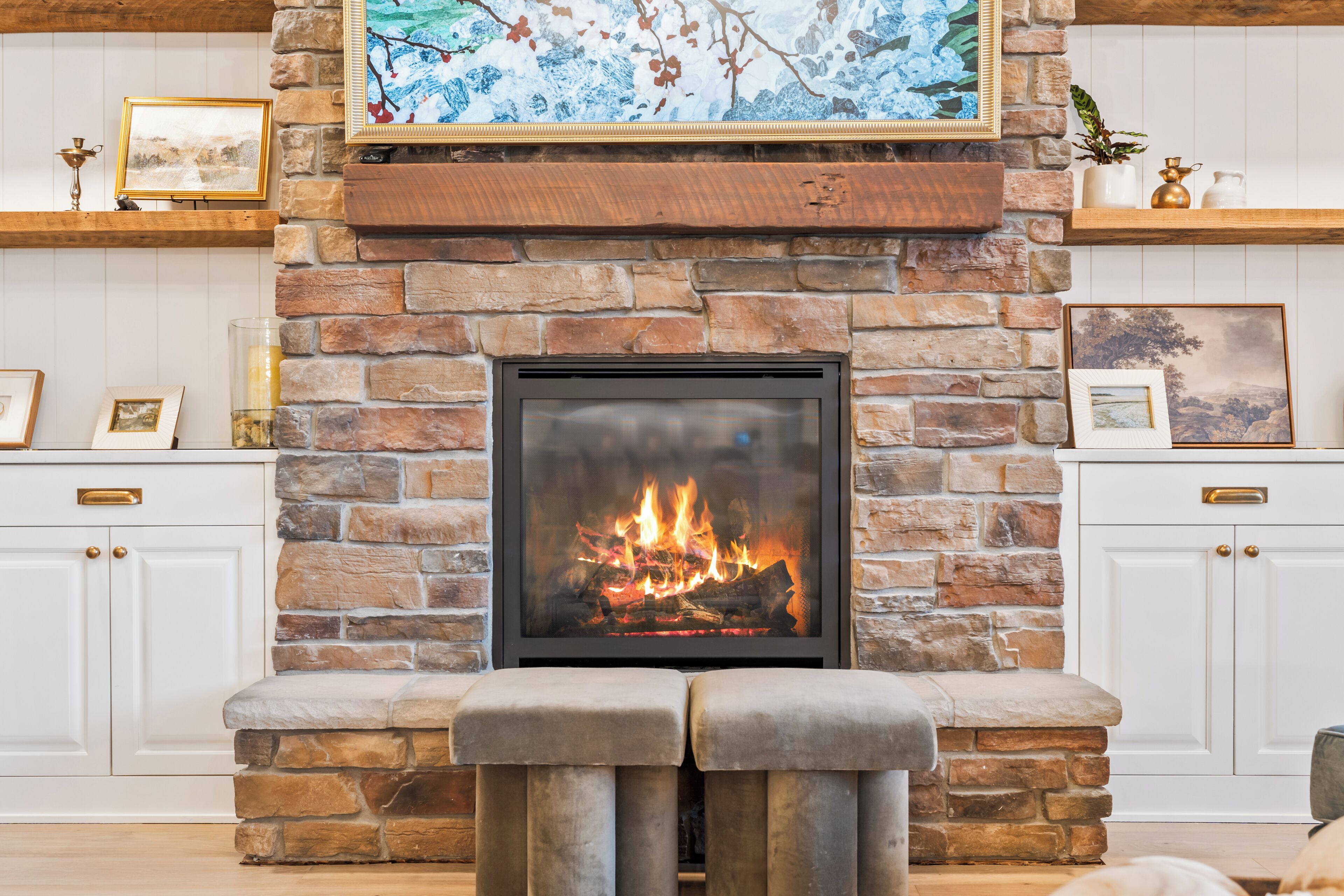 Une cheminée en pierre avec un feu, flanquée de meubles blancs et ornée de tableaux encadrés.