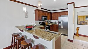 A modern kitchen with granite countertops, wooden cabinets, stainless steel appliances, and a central island with stools. - Remodeled Top-Floor Condo with Stunning Wrap-Around Balcony Offering Mountain, Ocean & Marina Views (Cabo Velas)