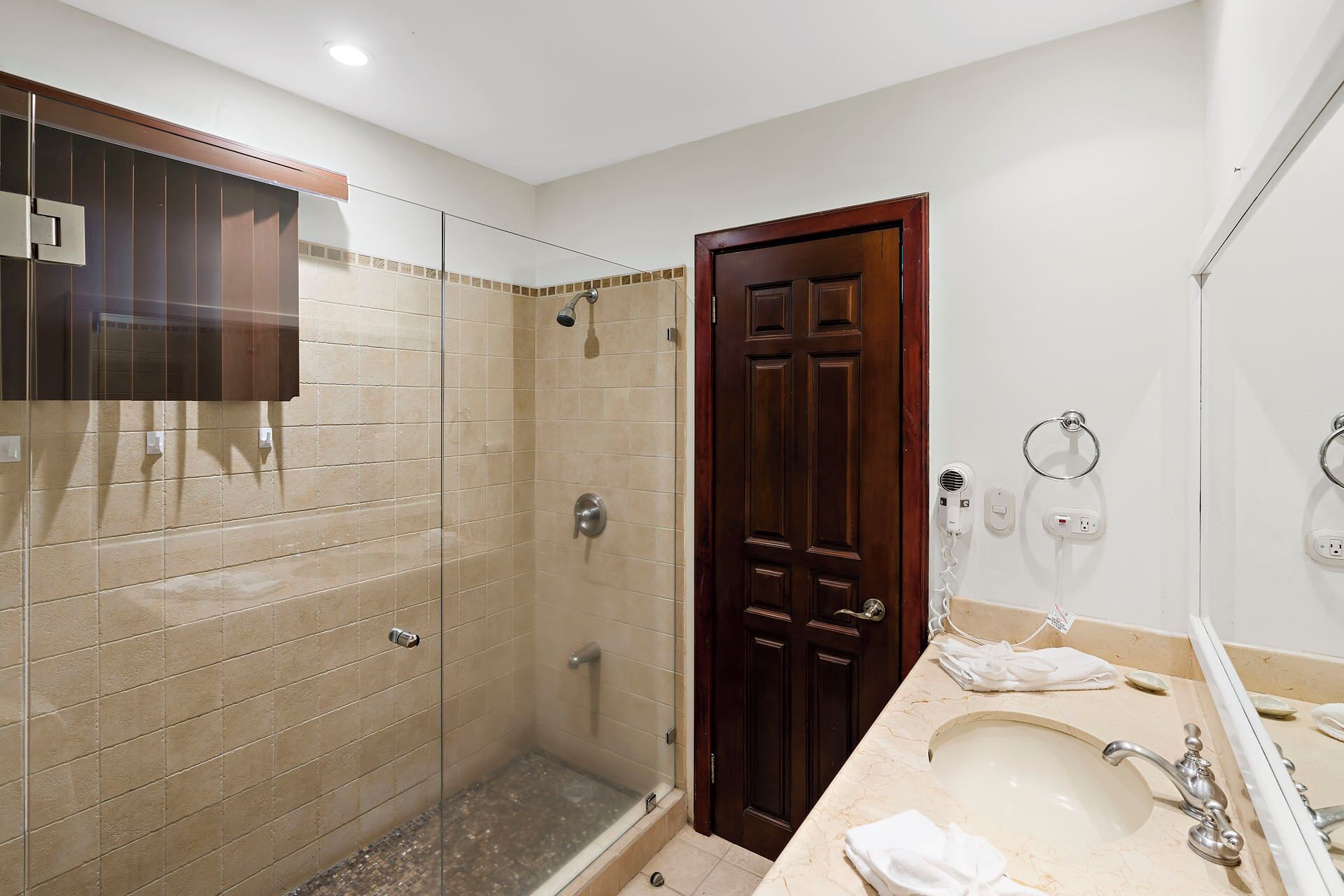 Un baño con cabina de ducha de vidrio, una puerta de madera, un lavabo con encimera de mármol y un espejo sobre el lavabo.