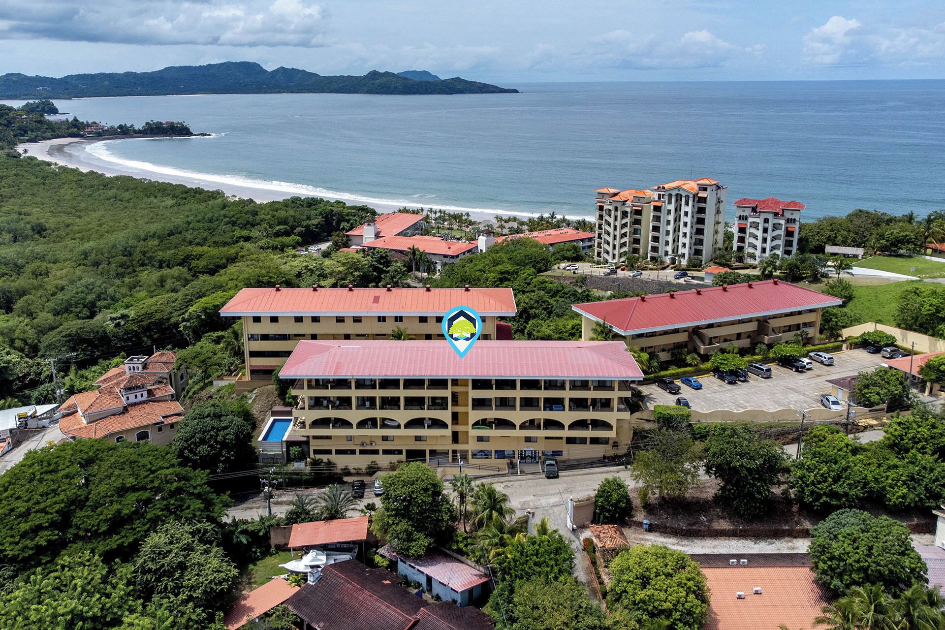 An aerial view of a coastal hotel with multiple buildings, a parking lot, and a beach in the background.