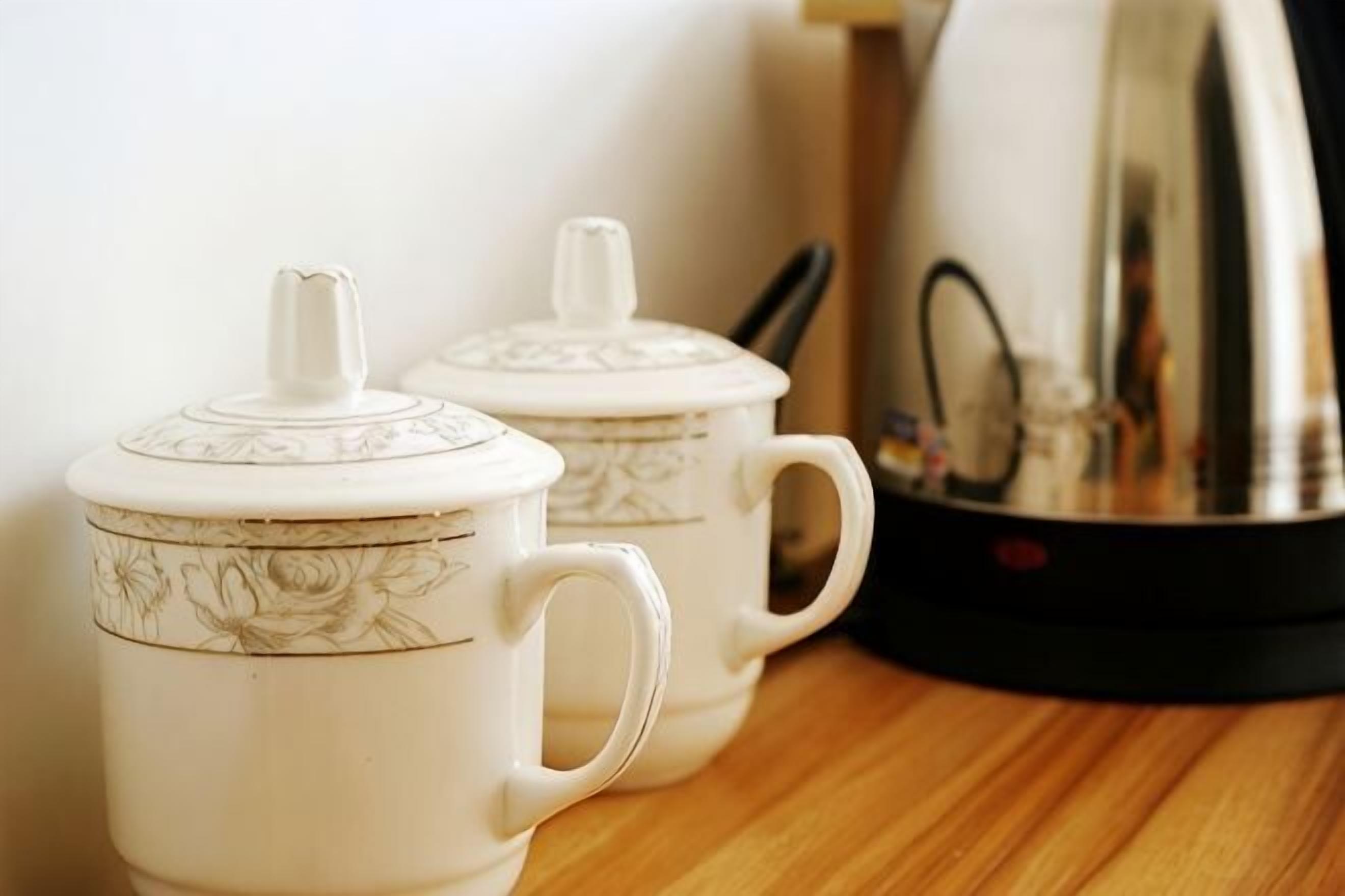 Two white ceramic cups with lids on a wooden surface, a black electric kettle, and a white wall in the background.