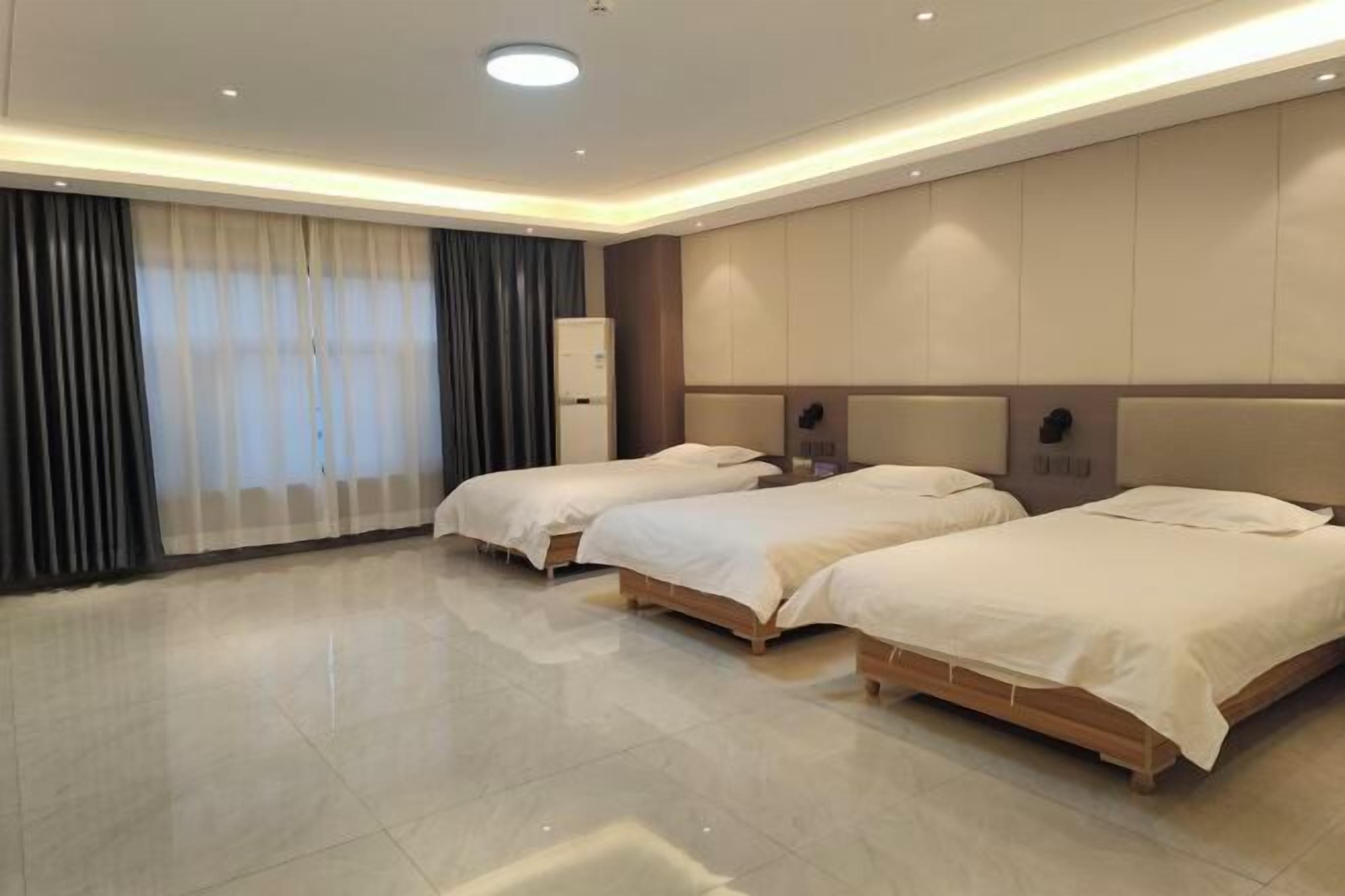 A hotel room with three beds, a large window with curtains, a minibar, and a ceiling light.