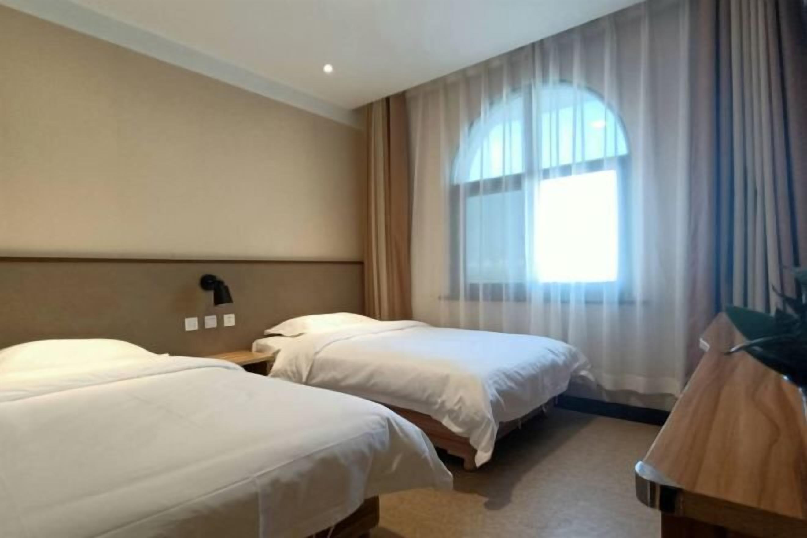 A hotel room with two beds, a nightstand, a lamp, and a large window with curtains.