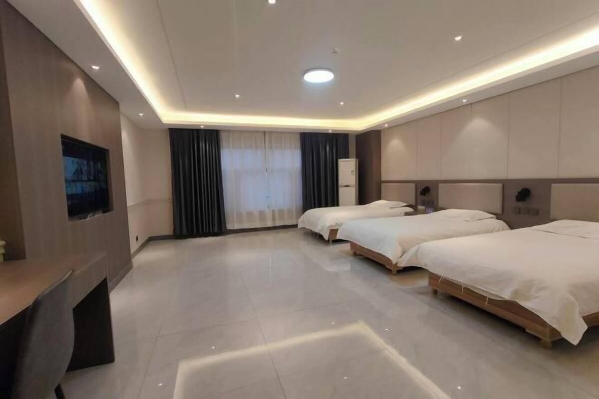 A modern hotel room with three beds, a flat-screen TV, and a large window with curtains.