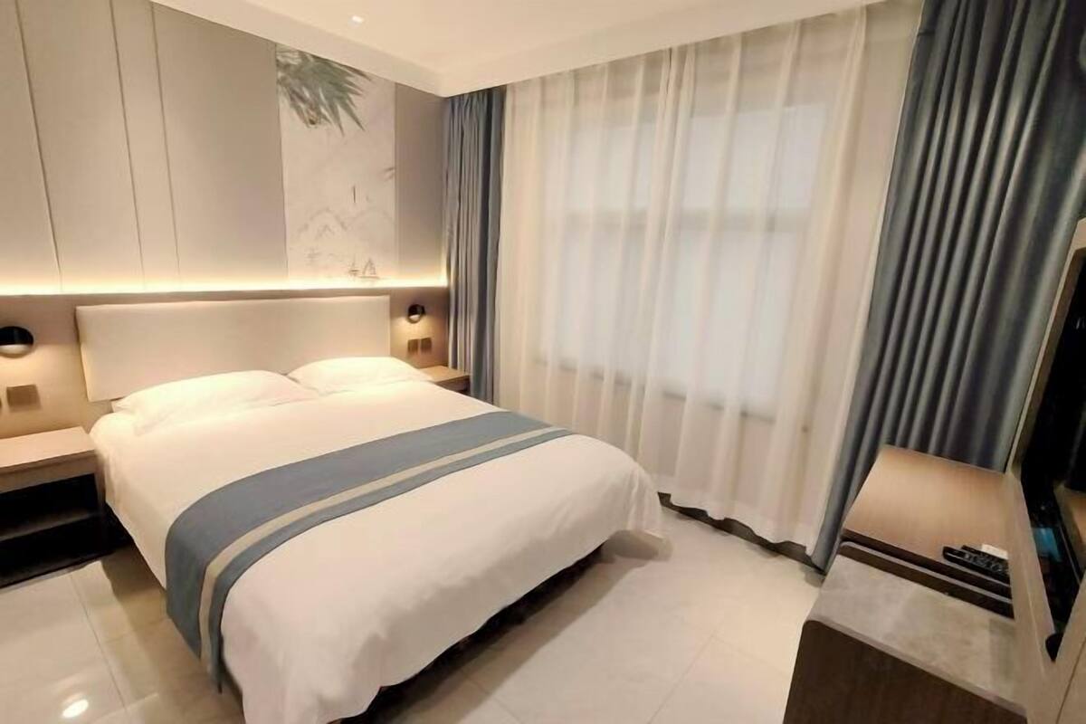 A hotel room with a bed, bedside tables, a TV, and a large window with curtains.