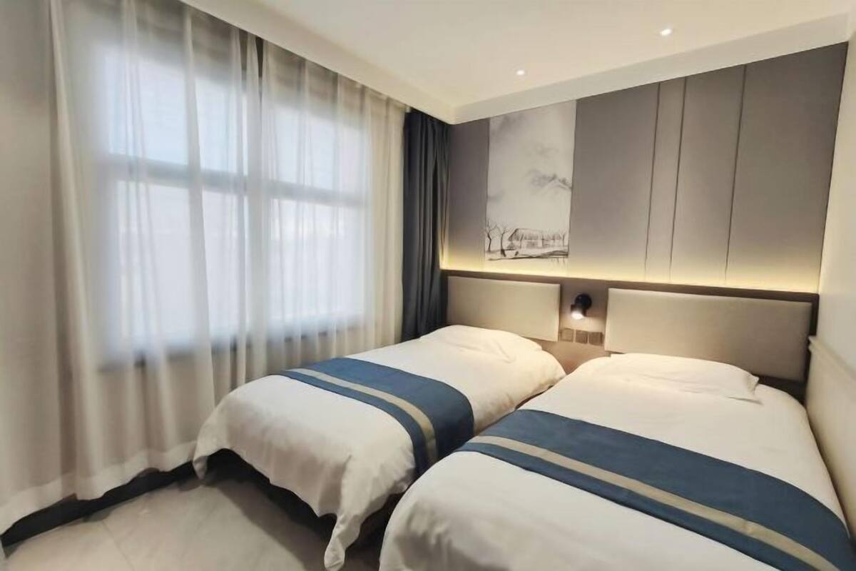 A hotel room with two beds, a large window with sheer curtains, a wall-mounted light, and a painting.