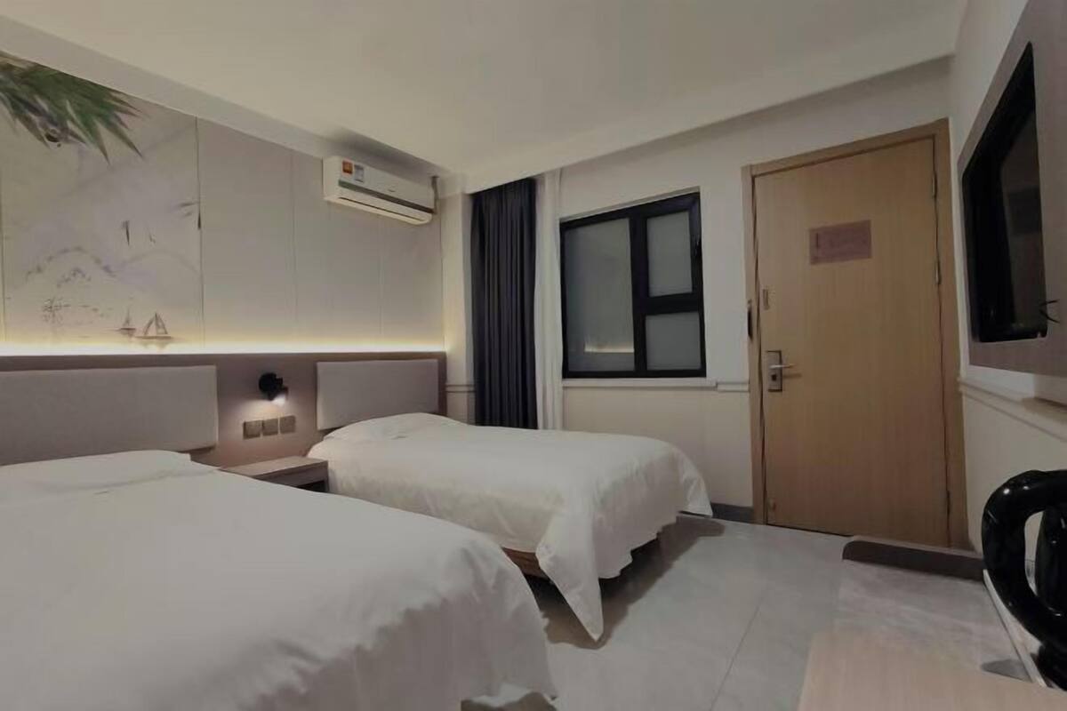 A hotel room with two beds, a wall-mounted air conditioner, a window with curtains, and a door.