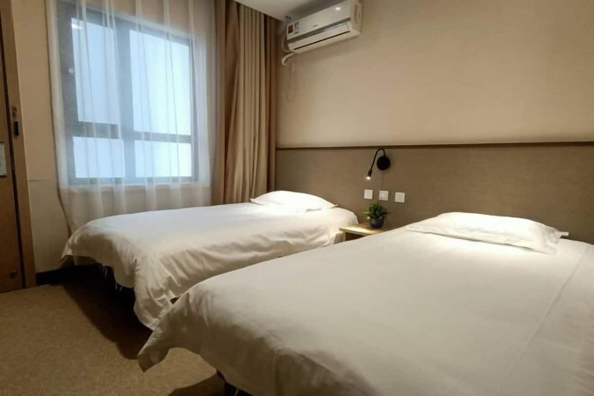 A hotel room with two beds, an air conditioning unit, a nightstand with a lamp, and a window with curtains.