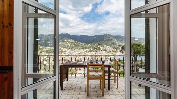 A balcony with a wooden table and chairs, offering a view of a mountainous landscape and a town below. - Casa Lucia a Rapallo (Rapallo)